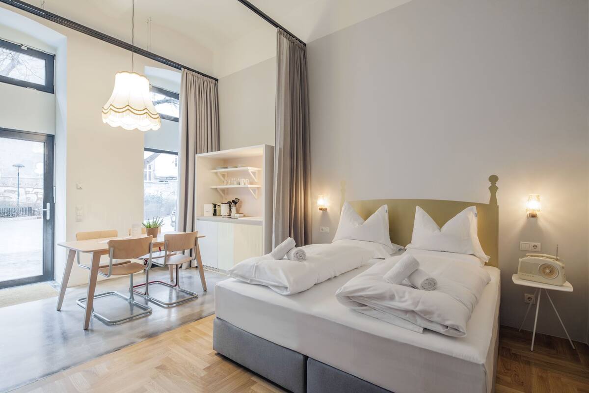 A modern hotel room with a large bed, a desk with chairs, a small table with a radio, and a view through the window.