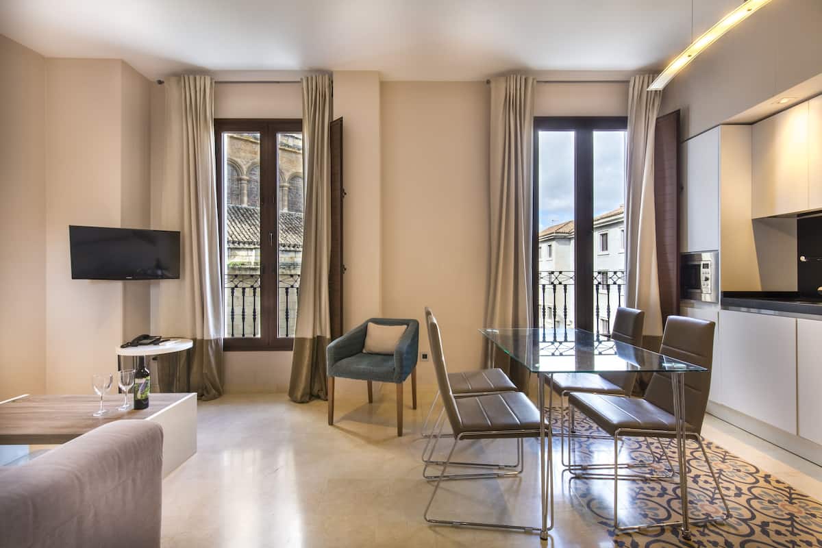 A modern hotel room with a glass dining table, comfortable seating, a flat-screen TV, and large windows offering a view of the cityscape.