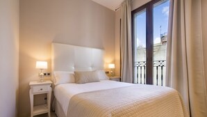 A neatly made bed with a white headboard, two bedside tables with lamps, and a view of a balcony through a large window with curtains. - Siloe Plaza (Granada)