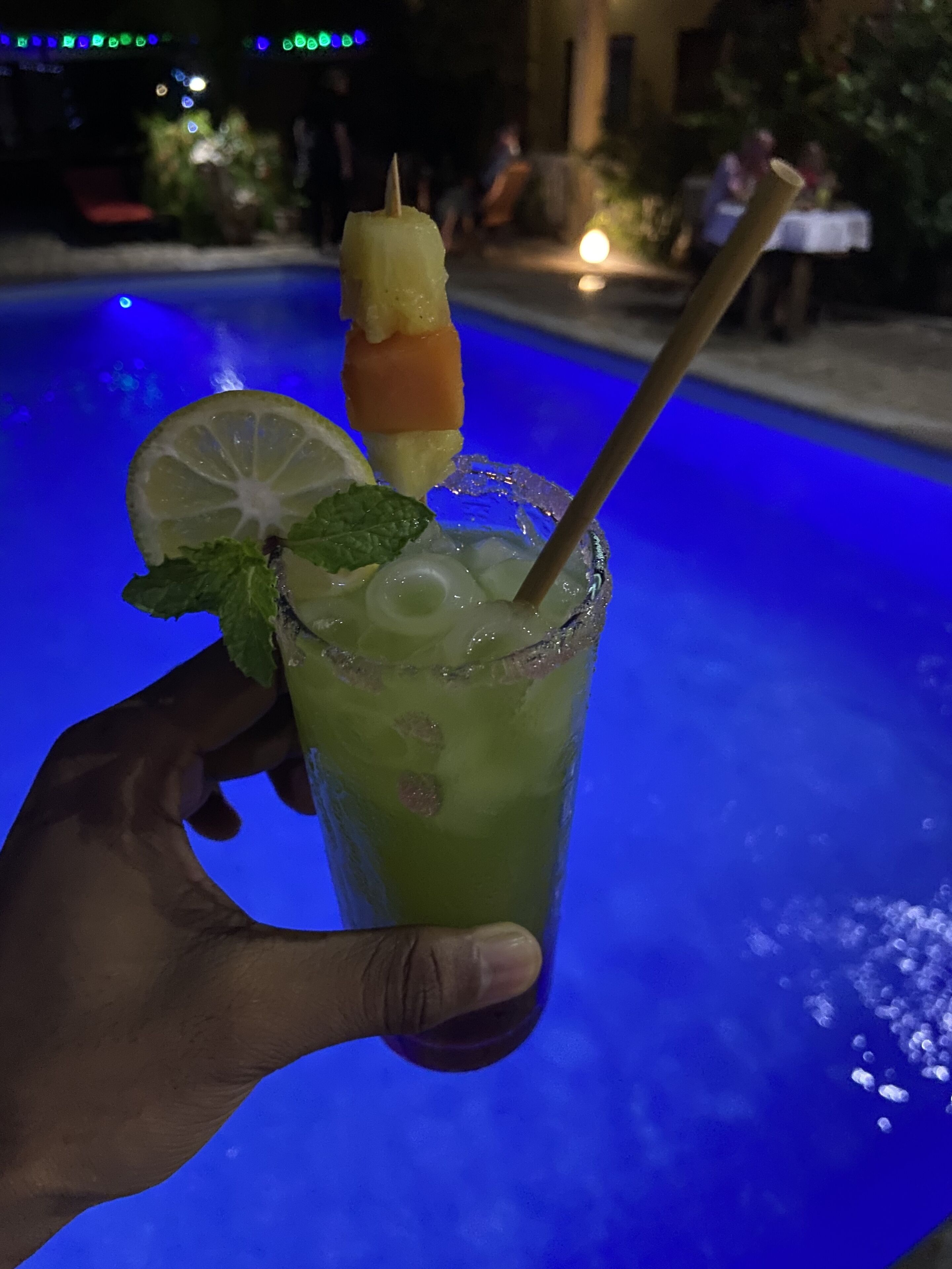 A hand holding a cocktail with a lime wedge and mint, with a pool in the background.