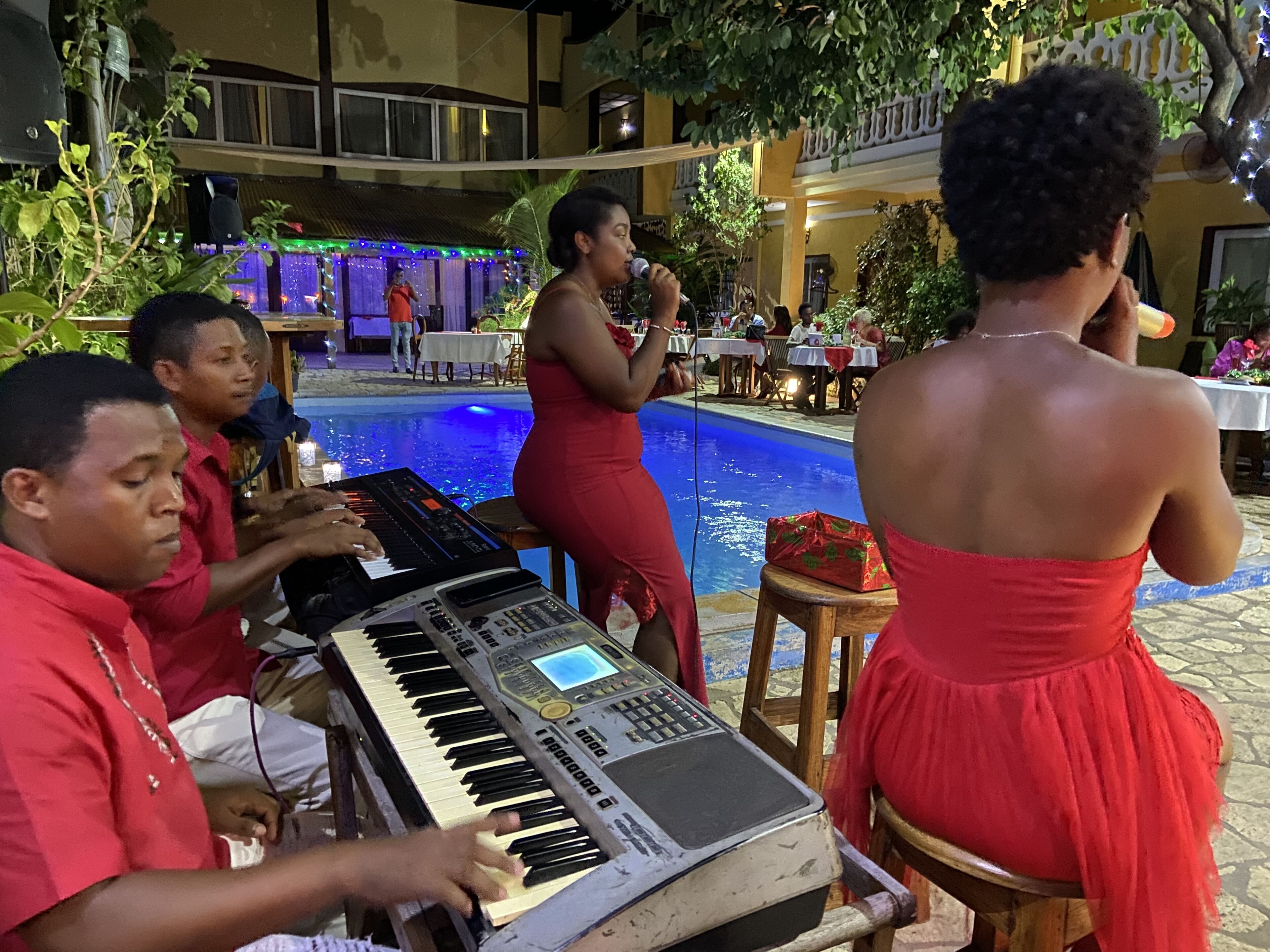 Un groupe de musique joue en direct au bord d’une piscine, et une femme vêtue d’une robe rouge est assise à proximité.
