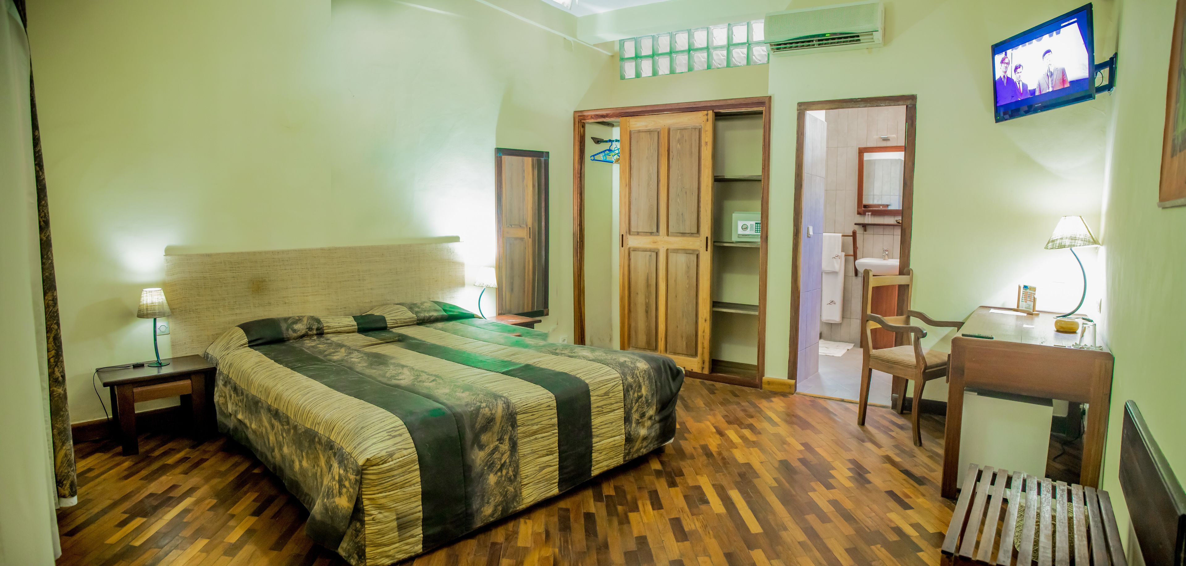 Un dormitorio con una cama de madera, una mesita de noche con lámpara, un armario, una pequeña mesa con una silla y un televisor montado en la pared.