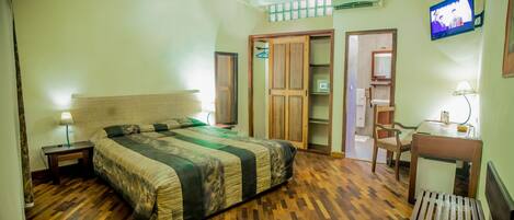 Un dormitorio con una cama de madera, una mesita de noche con lámpara, un armario, una pequeña mesa con una silla y un televisor montado en la pared.