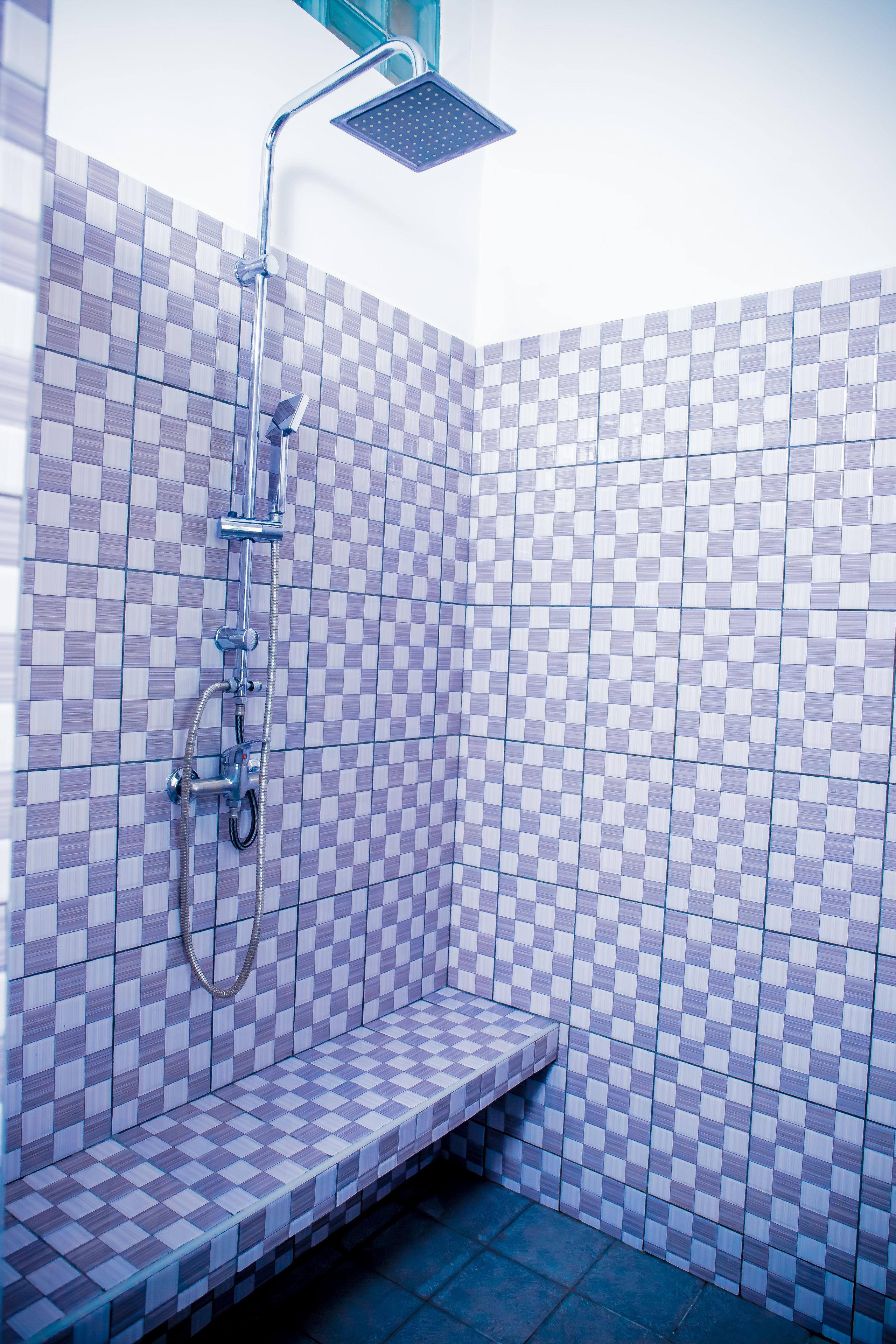 A bathroom with checkered tile walls, a shower with a handheld showerhead, and a bench.