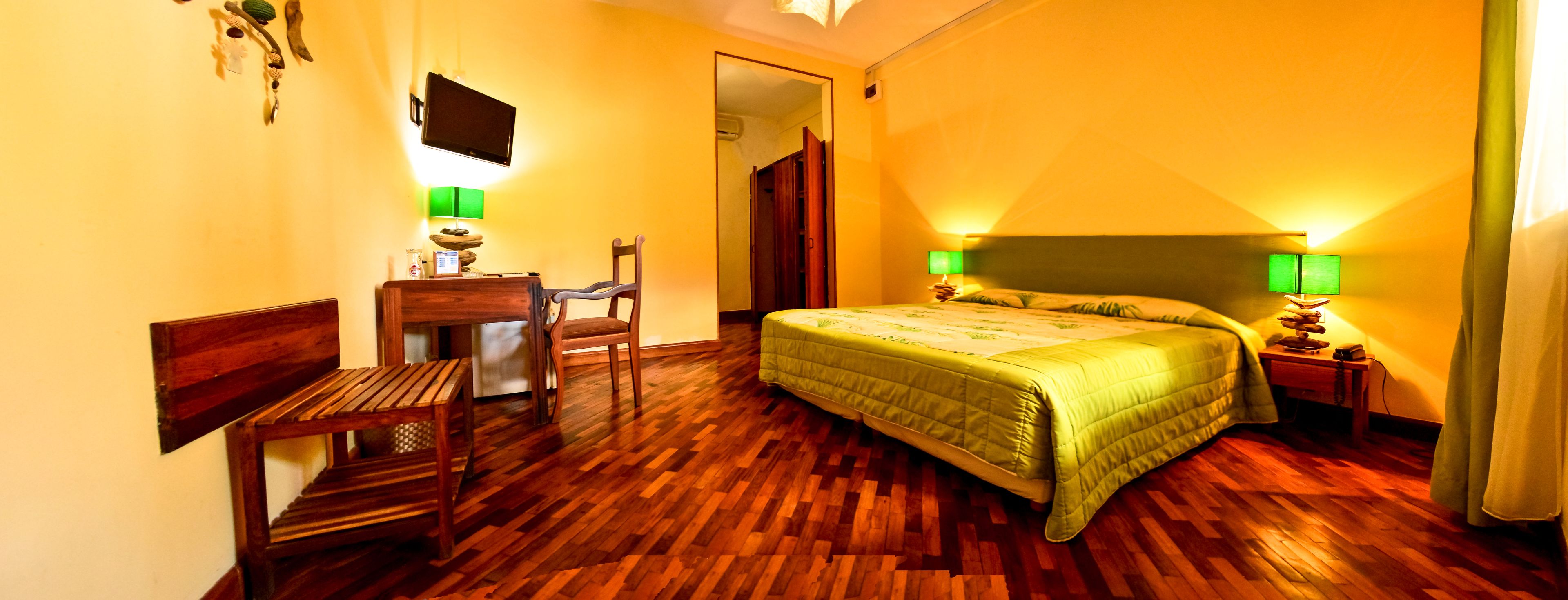 Habitación de hotel con piso de madera, cama con colcha verde, escritorio con silla, televisor montado en la pared y dos lámparas de noche.