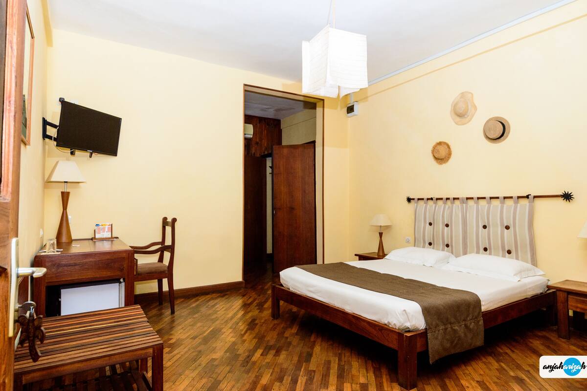 Un dormitorio con una cama de madera, un escritorio con una silla, un televisor montado en la pared y una puerta que da a otra habitación.