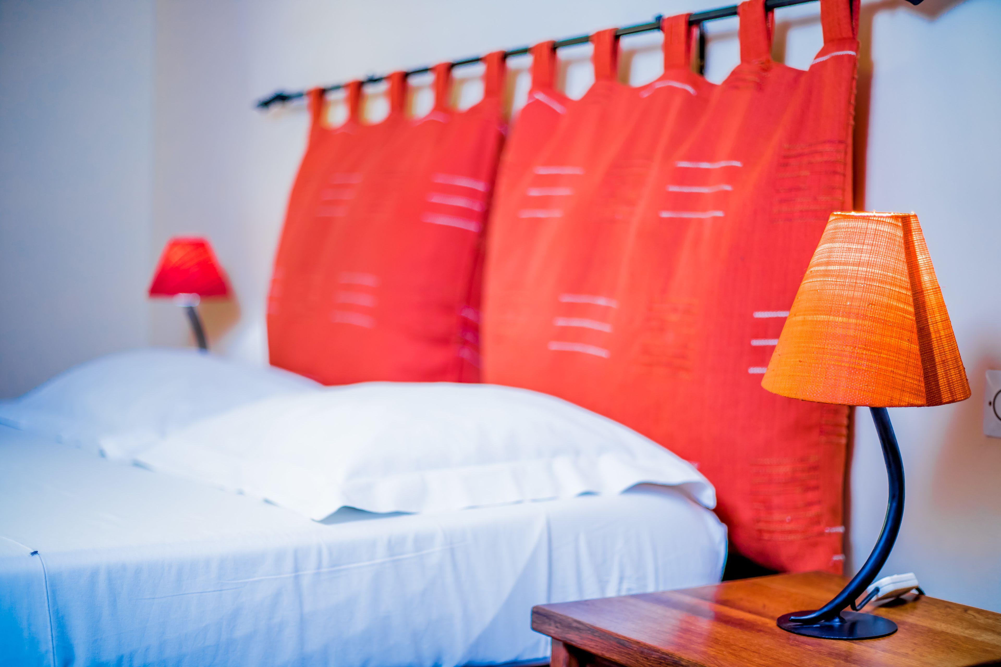 Een bed met witte lakens, een rood gordijn en een tafellamp op een houten nachtkastje.