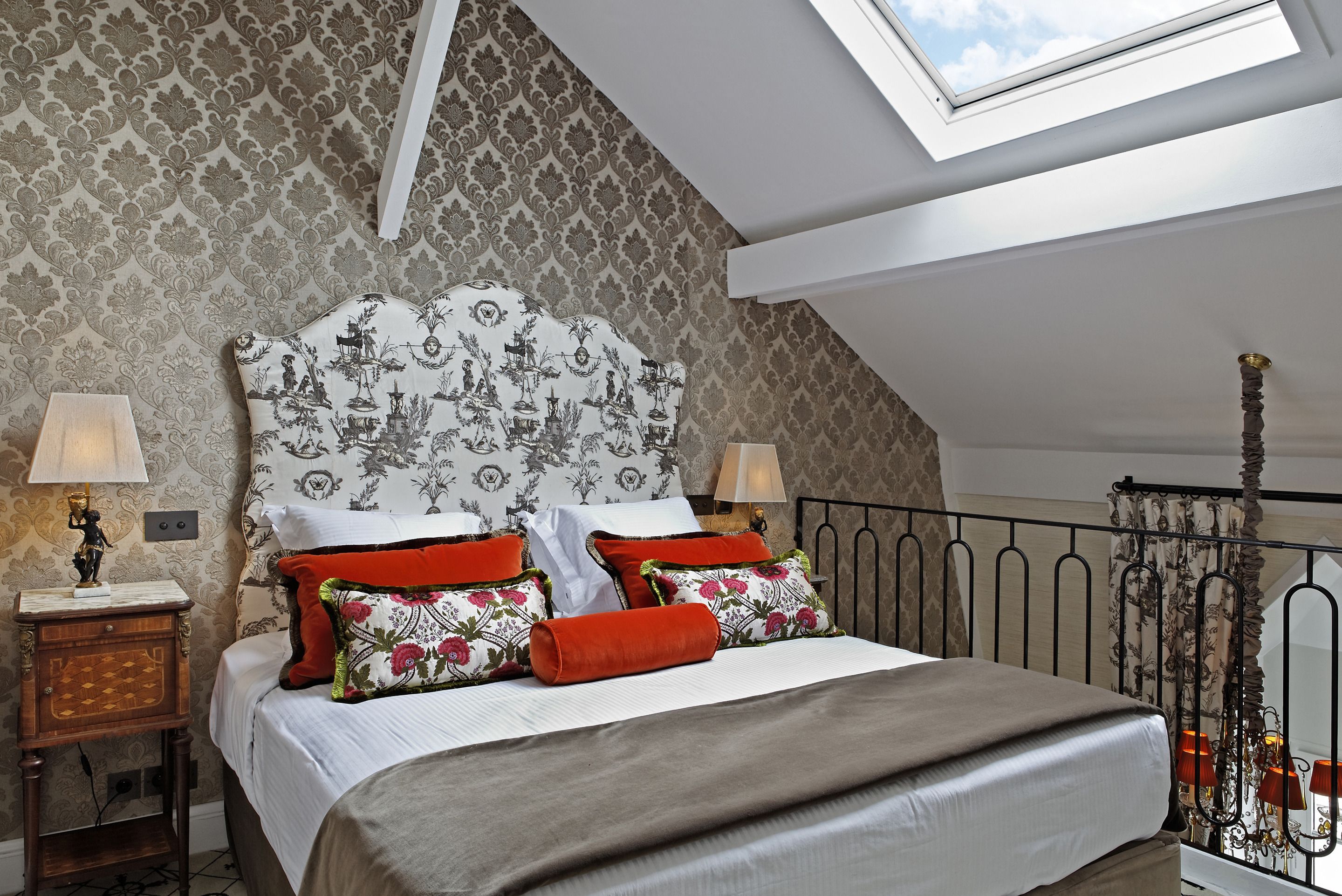 Une chambre avec un plafond haut, un papier peint à motifs, un puits de lumière, un lit avec de nombreux oreillers, une table de chevet avec une lampe et une balustrade en métal.