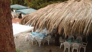 Un área de comedor al aire libre con sombrillas de techo de paja, sillas de plástico blanco y mesas dispuestas para los comensales.
