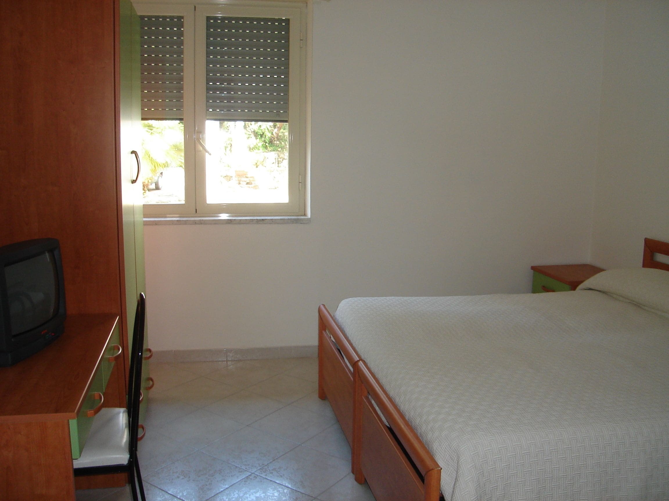 A bedroom with a bed, a television, a window with blinds, and a small table.