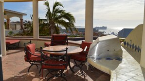 Eine Terrasse im Freien mit einem runden Tisch und Stühlen, einem Grill und Blick auf das Meer und die Skyline der Stadt.