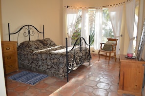 A bedroom with a metal bed frame, a wooden dresser, a chair, and a window with curtains. - Casa Bahia (Cabo San Lucas)