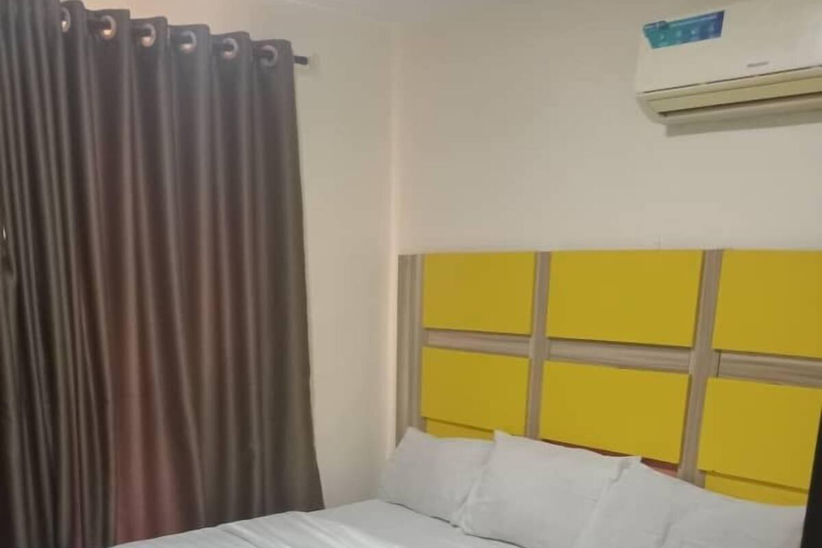 Uma cama bem arrumada com roupa de cama branca, uma cabeceira com painel amarelo e uma janela com cortinas.
