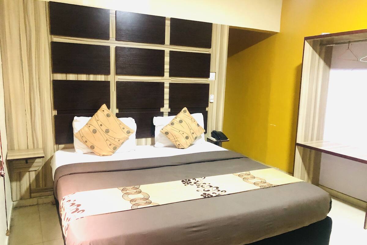 Quarto de hotel com uma cama grande, cabeceira com janela, mesa de cabeceira e pequena escrivaninha com cadeira.