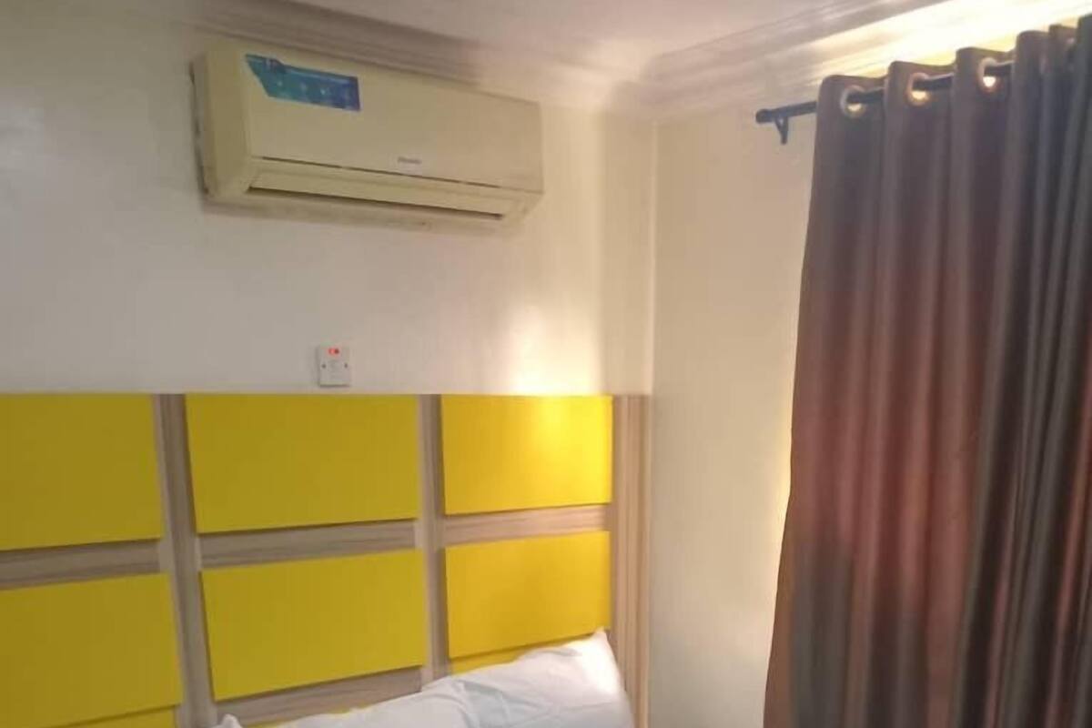 Quarto de hotel com cama, cabeceira com painéis amarelos, aparelho de ar condicionado e cortina.