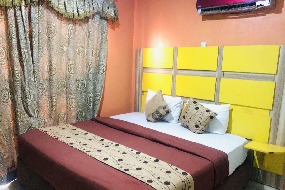 Uma cama com colcha vermelha e manta estampada, cabeceira com design em painel amarelo e cortina com padrão floral.