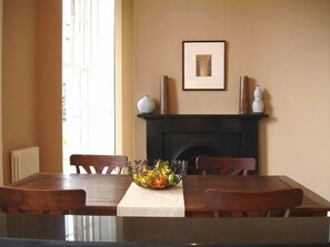 Uma sala de jantar com uma mesa de madeira, cadeiras, toalha de mesa branca, uma fruteira e uma lareira com vasos decorativos.