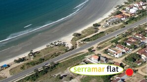 An aerial view of a coastal area with a beach, a small island, and a residential area with houses and roads. - Serramar Flat (Ubatuba)