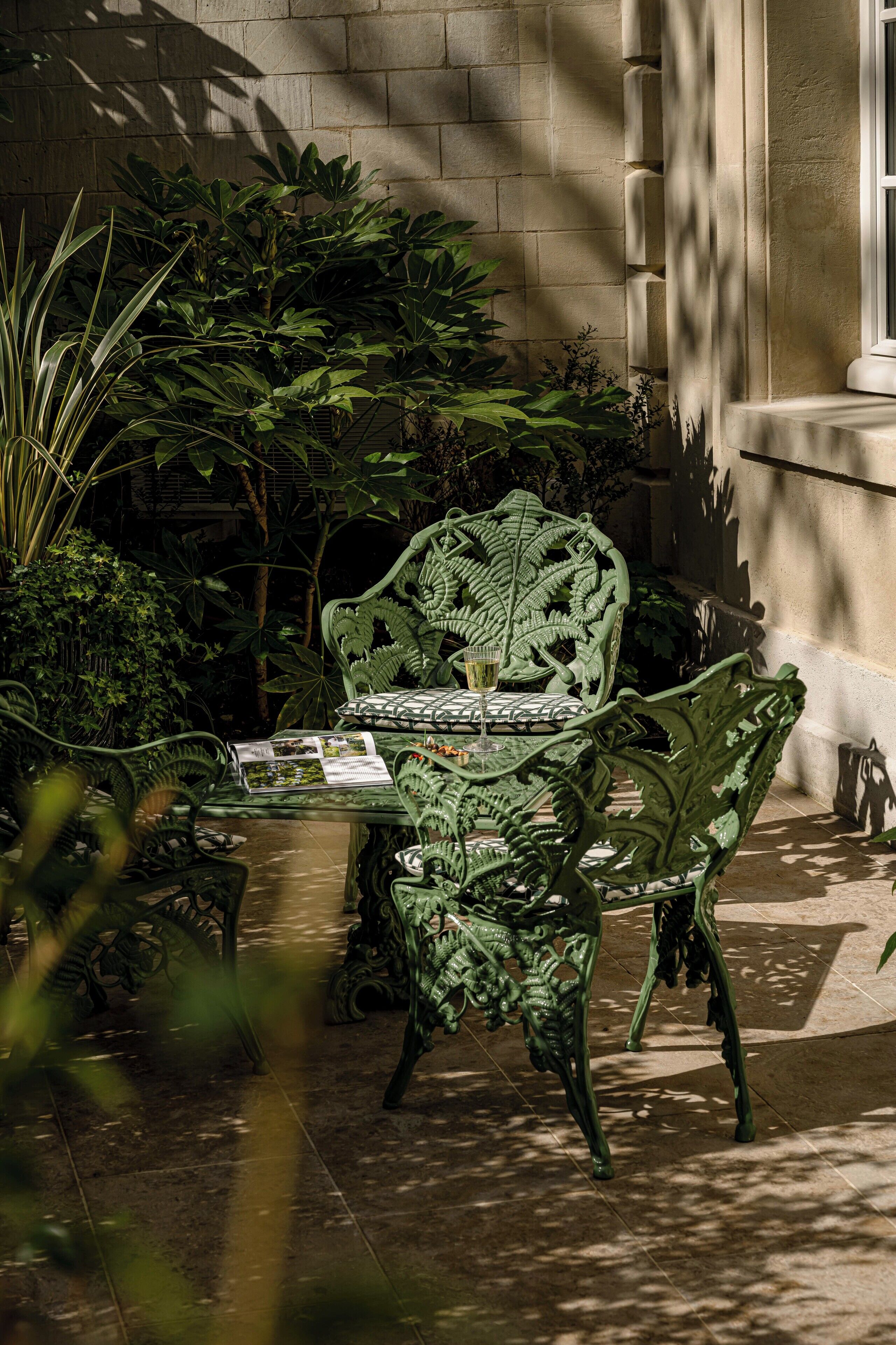 A green wrought iron table and chairs set in a shaded outdoor area with a tiled floor, surrounded by lush greenery and a stone wall.