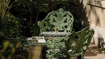 A green wrought iron table and chairs set in a shaded outdoor area with a tiled floor, surrounded by lush greenery and a stone wall.