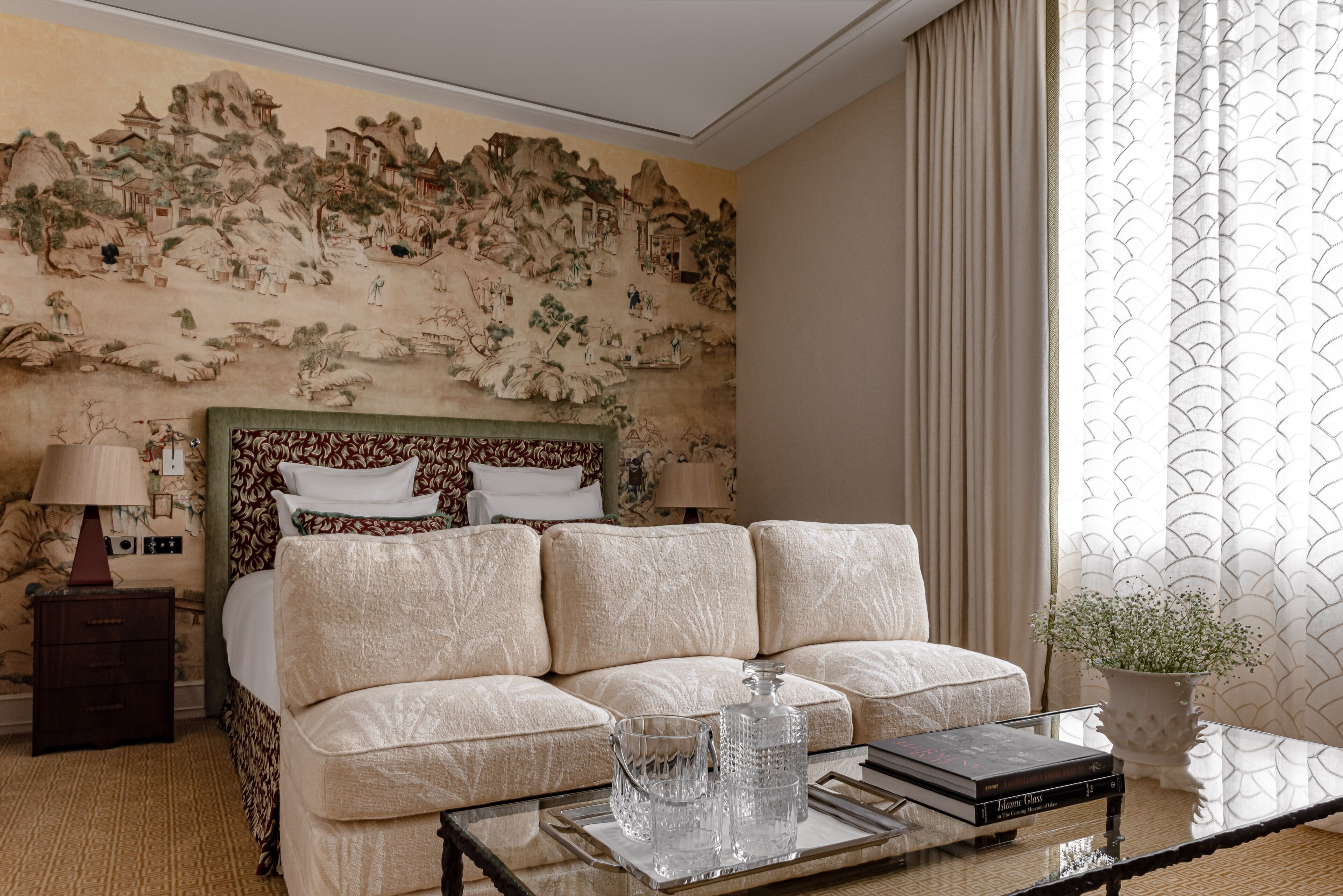 A hotel room with a traditional mural, a sofa bed, a bedside table with lamps, and a glass coffee table with books and decorative items.