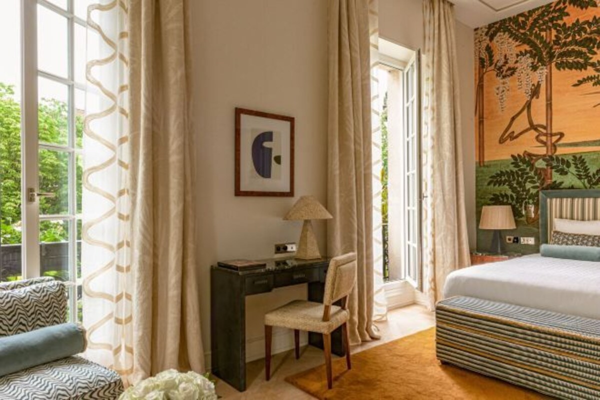 Un dormitorio con cama, escritorio y silla. Hay una ventana con cortinas, una pared con un cuadro, y vistas de vegetación al exterior.