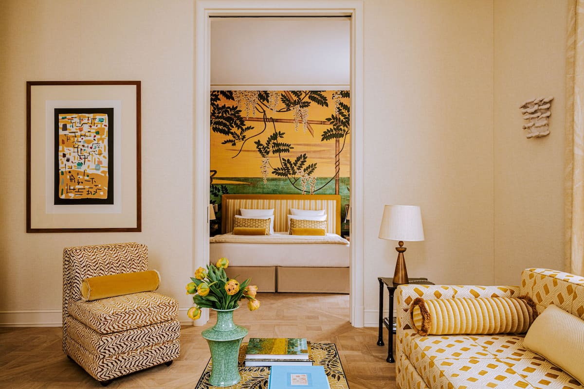 Una habitación con un sillón estampado, un sofá, una mesita de noche con lámpara y una cama con ropa de cama amarilla.