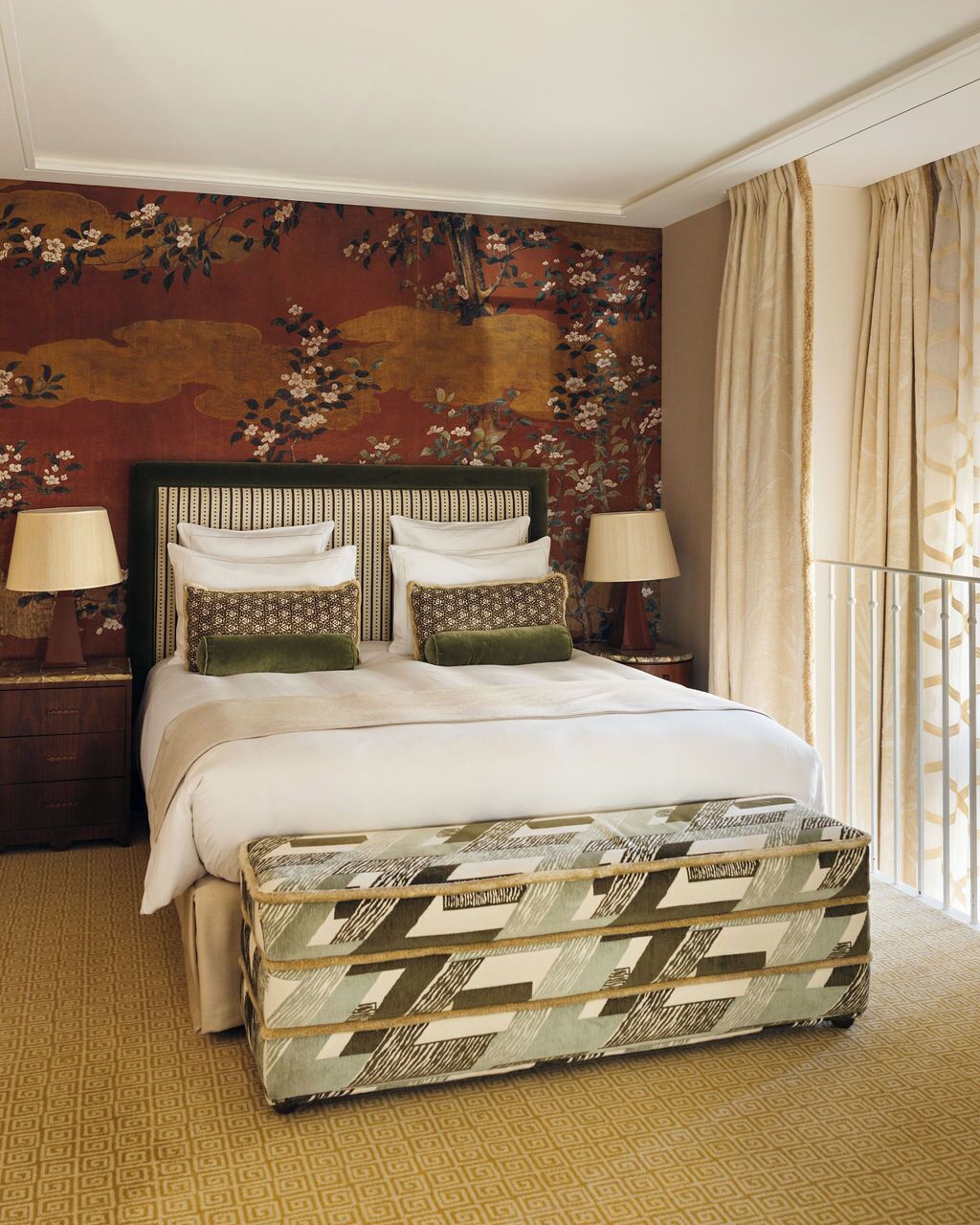 Habitación de hotel con una cama grande, dos mesitas de noche con lámparas, un reposapiés estampado, papel tapiz floral y una ventana con cortinas.