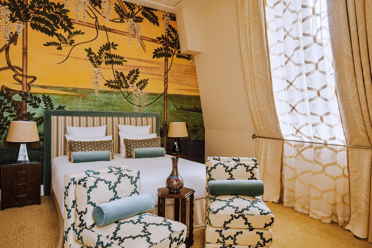 Una habitación de hotel con una cama grande, dos mesitas de noche, una silla, una pequeña mesa con un florero y una ventana con cortinas.