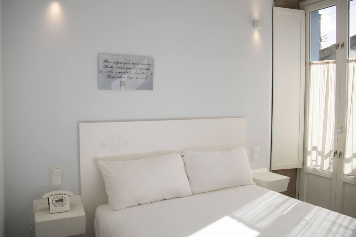 Une chambre à coucher avec un lit, une table de chevet avec un téléphone, une lampe fixée au mur et une fenêtre avec des rideaux.