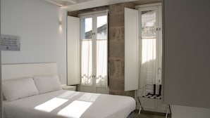 Ein Schlafzimmer mit Bett, einem Fenster mit Vorhängen, einem Sessel und einem an der Wand befestigten Regal.