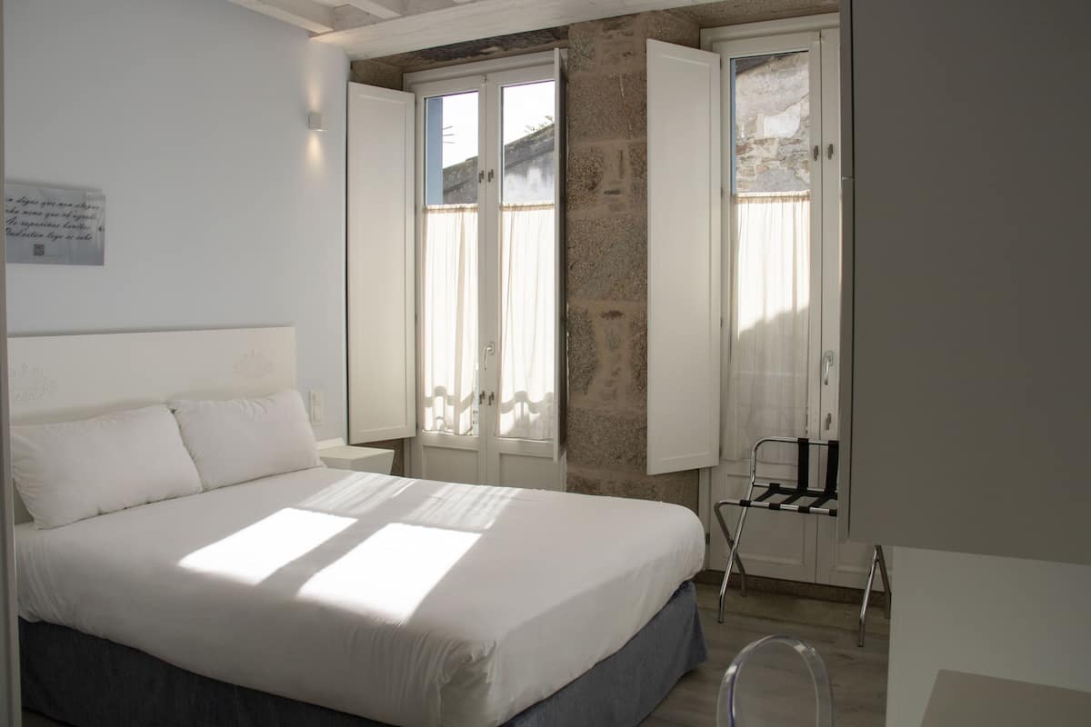 Une chambre à coucher avec un lit, une fenêtre avec des rideaux, une chaise et une étagère fixée au mur.