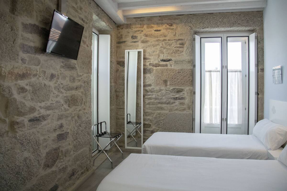 Une pièce aux murs en pierre, un téléviseur à écran plat fixé au mur, un grand miroir, deux lits avec du linge de lit blanc et une fenêtre avec des rideaux.
