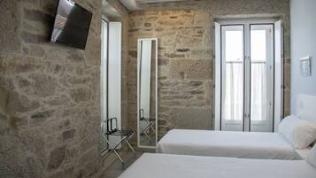 A room with stone walls, a flat-screen TV mounted on the wall, a large mirror, two beds with white bedding, and a window with curtains.