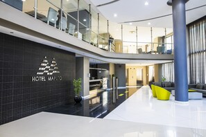A modern hotel lobby with a reception desk, seating area, and a staircase leading to an upper level.