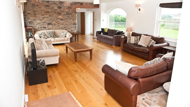 A spacious living room with a stone wall, wooden flooring, and multiple seating areas including a brown leather sofa and a beige sofa. - The Mill House (Earlston)