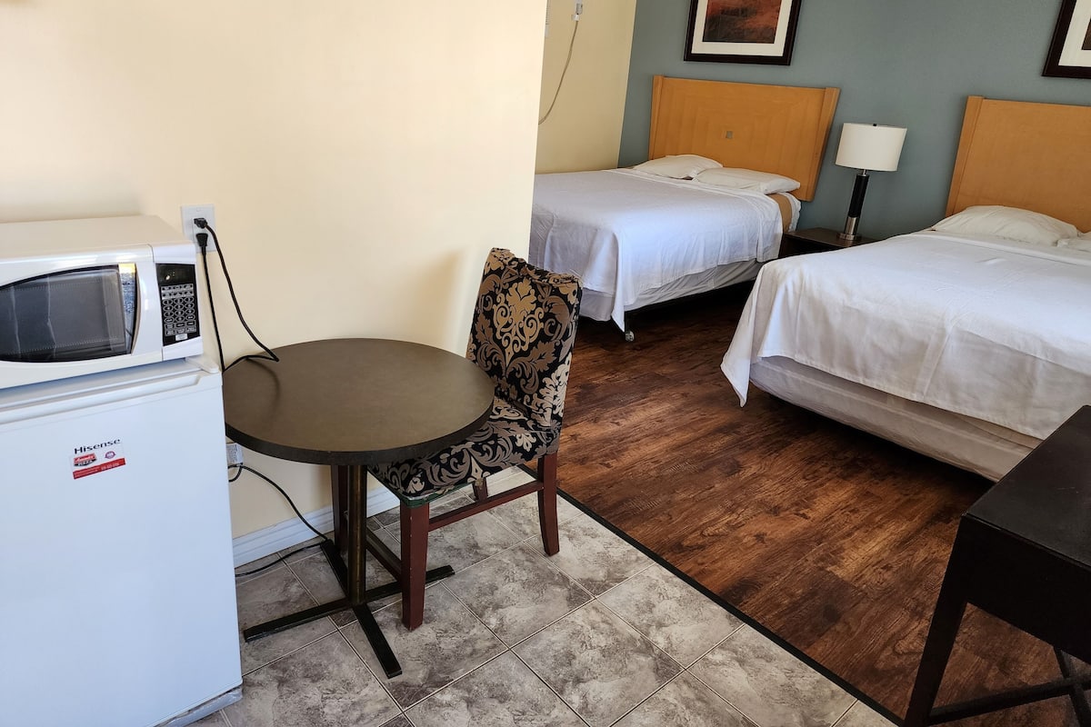 A hotel room with a microwave, a small round table, two beds, a chair, and a nightstand with a lamp.