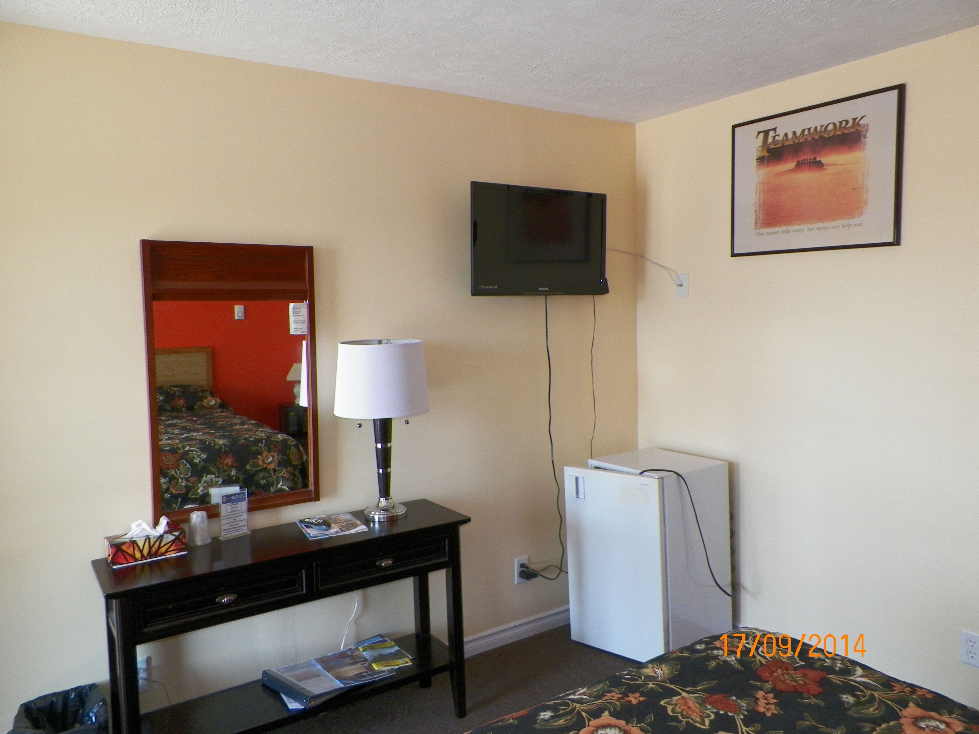 A hotel room with a bed, a desk with a lamp, a television on the wall, and a refrigerator.