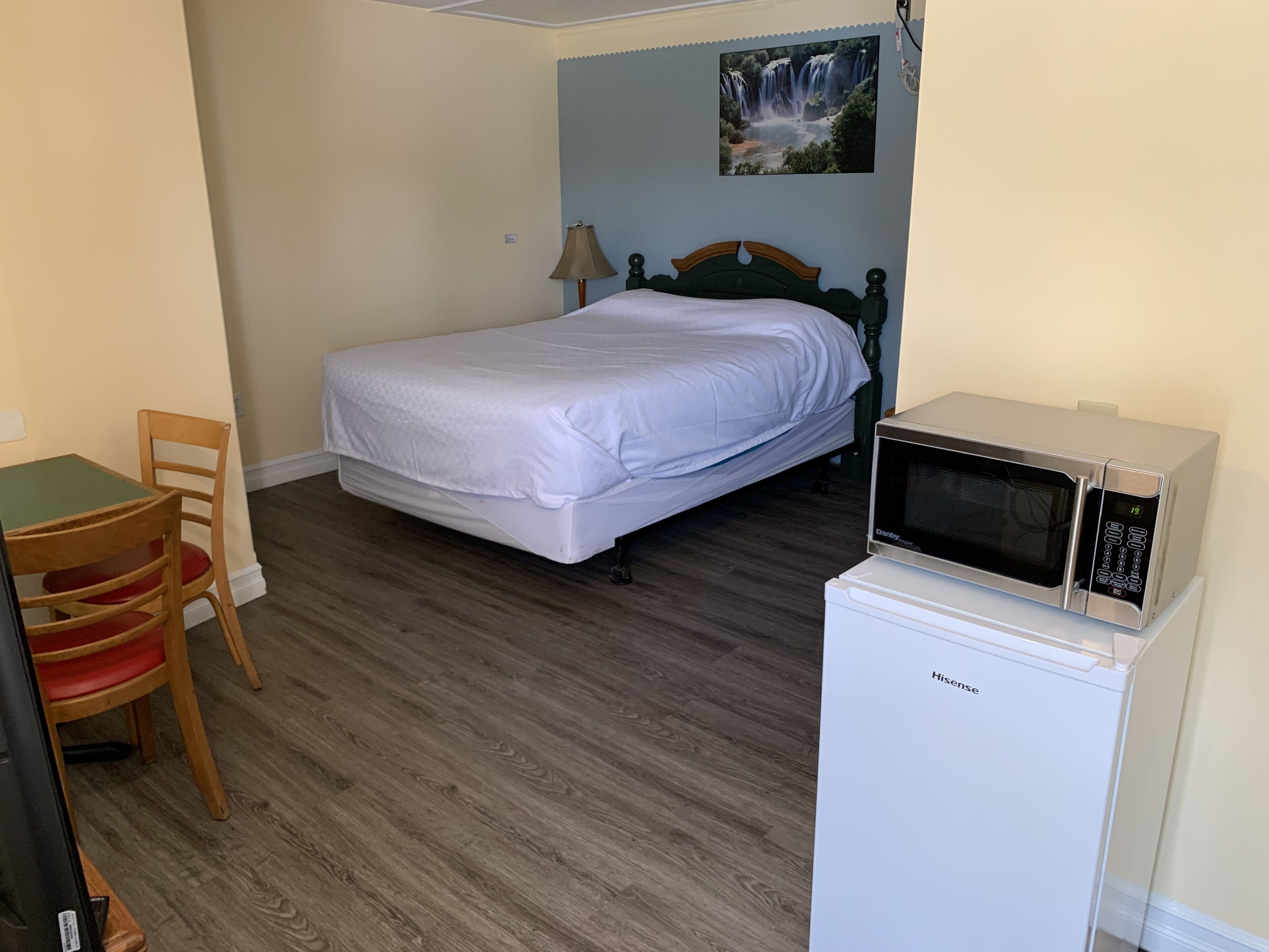 Una habitación pequeña y ordenada con cama, escritorio, silla, microondas y un cuadro de una cascada en la pared.