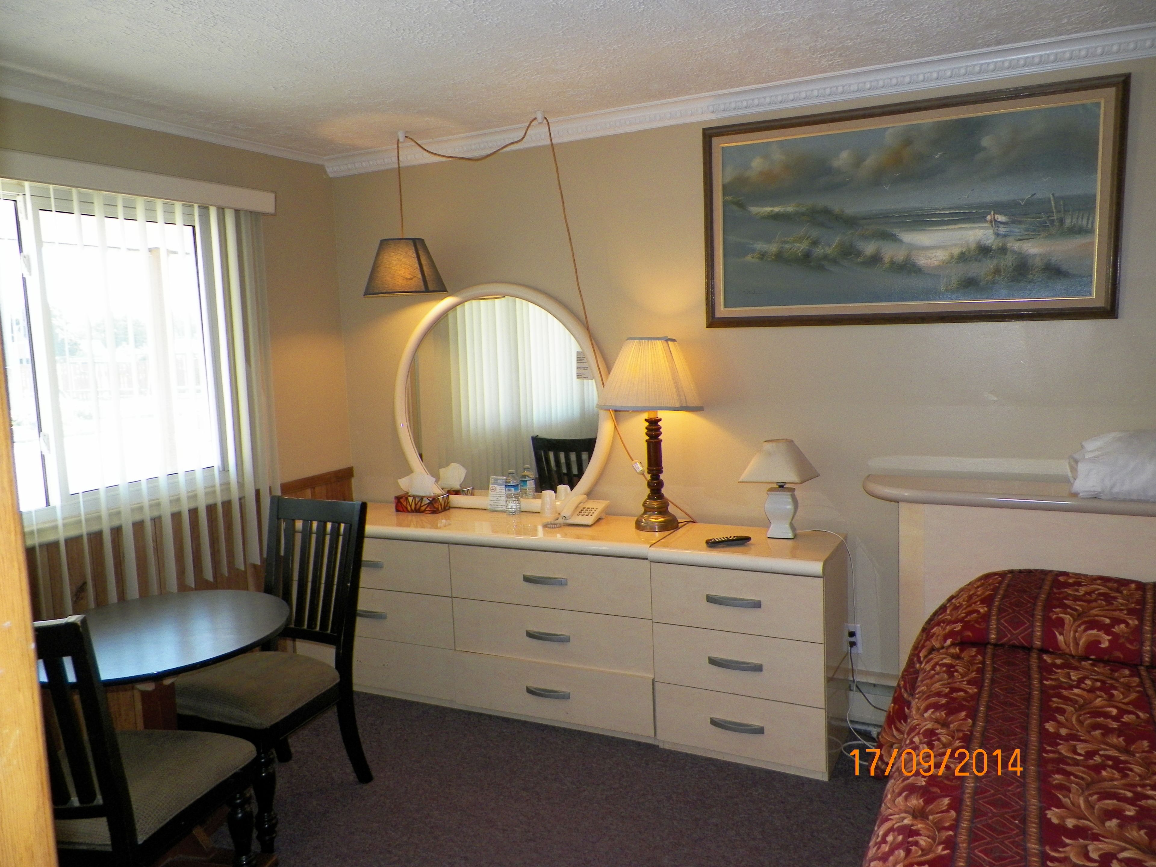 A hotel room with a desk, chair, dresser, lamps, and a painting on the wall.