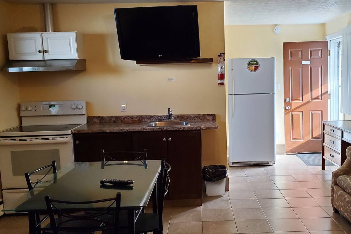 A compact kitchen with a stove, sink, and refrigerator, a dining table with chairs, and a sofa.