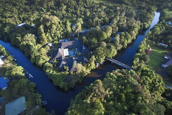An aerial view of a riverside property with a house surrounded by dense trees, a bridge, and a small dock.