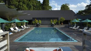 A rectangular pool with a tiled floor, surrounded by lounge chairs and umbrellas, with a wooden deck and a dark building in the background. - Mulu Marriott Resort (Mulu)
