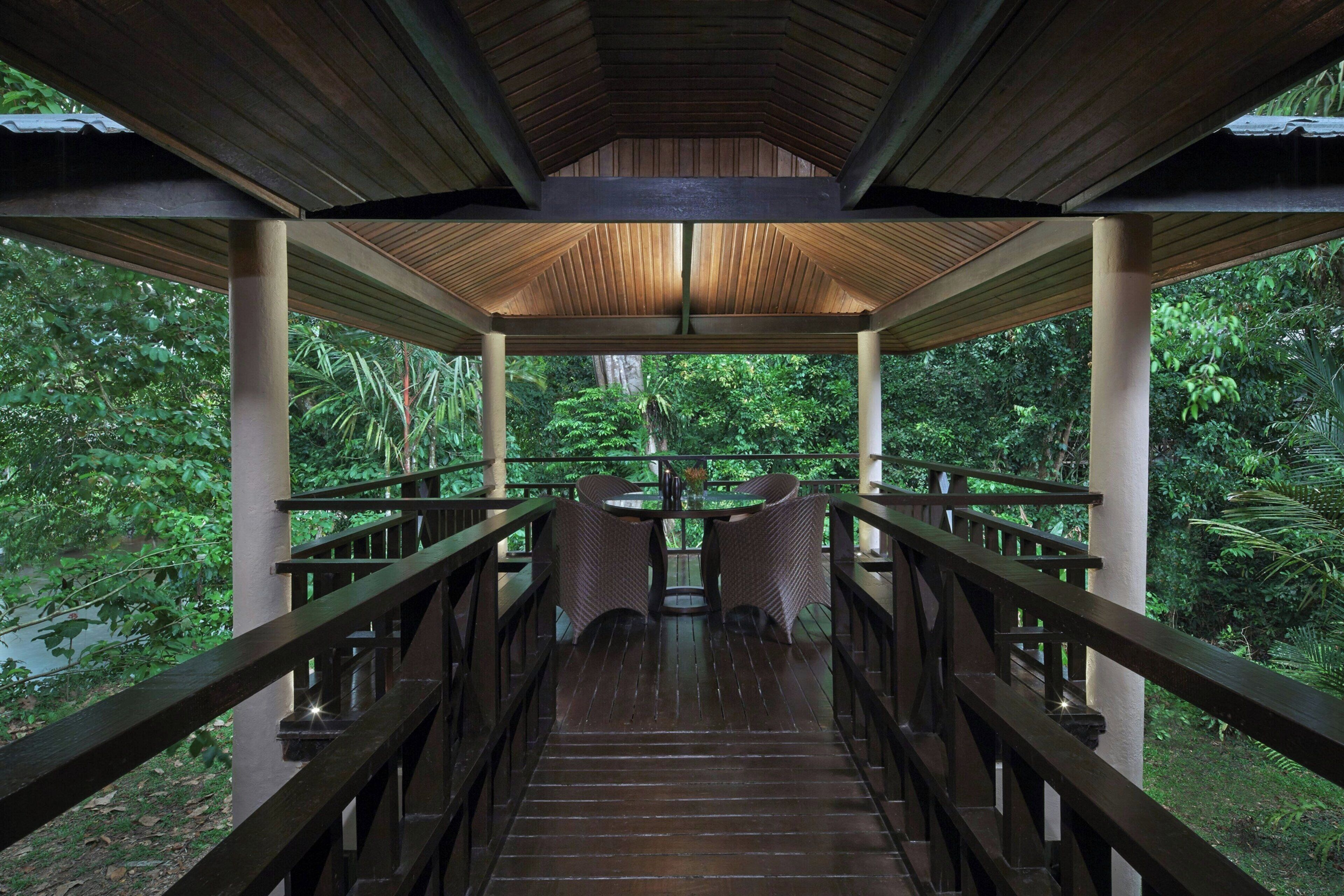 A covered wooden walkway with railings, leading to a table with chairs, surrounded by lush greenery.