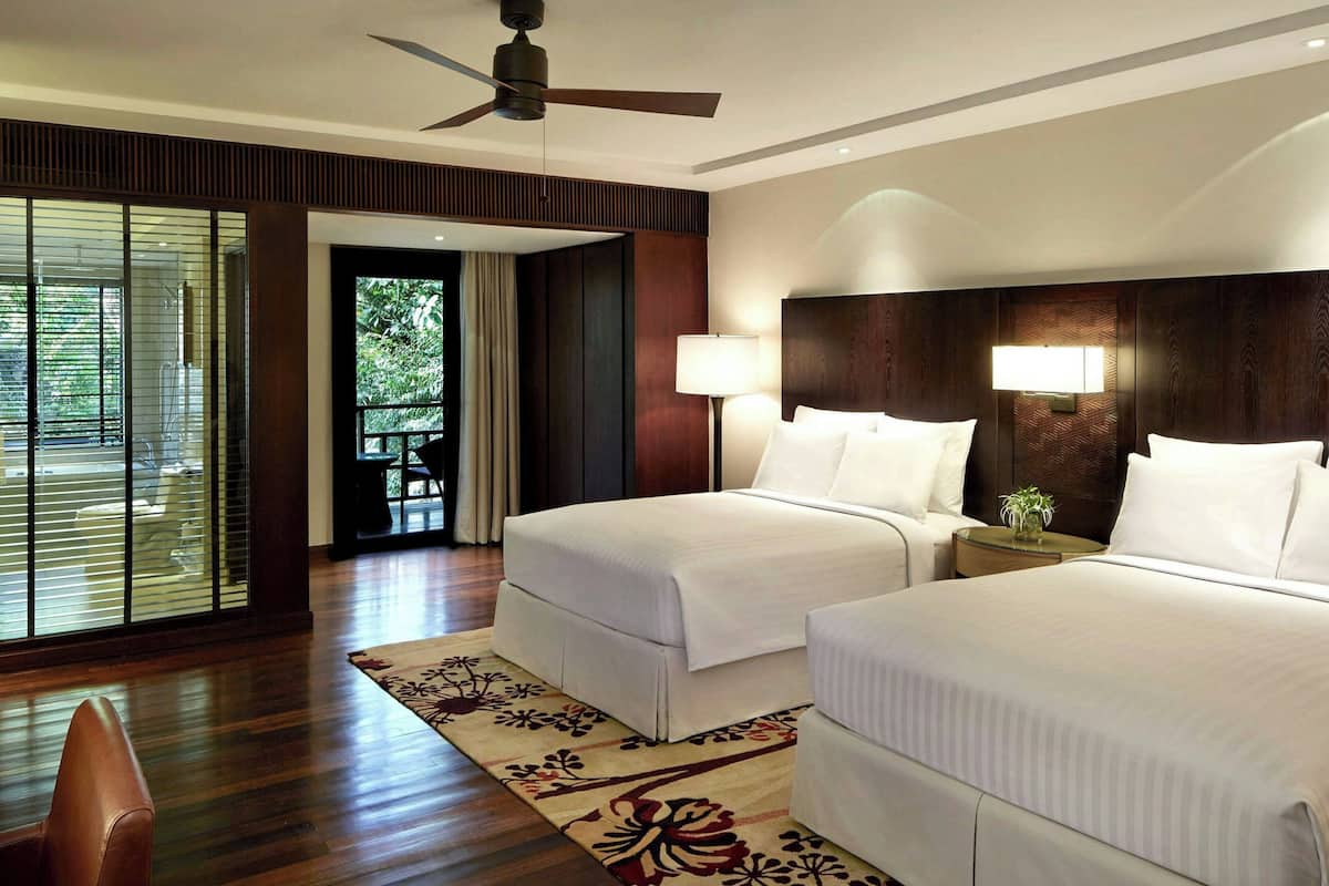 A hotel room with two beds, a ceiling fan, a nightstand with a lamp, a window with blinds, and a wooden floor.