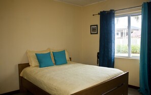 A single bed with a wooden frame and a white bedspread, two blue pillows, a small bedside table, a telephone, and a window with blue curtains. - Quest Lodge (Accra)