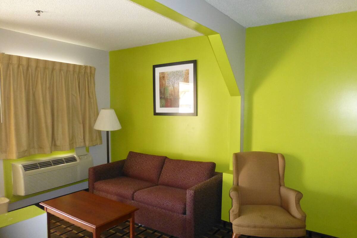 A room with a green accent wall, a brown sofa, a wooden table, a beige armchair, a framed picture, a window with beige curtains, and a floor with a checkered pattern.