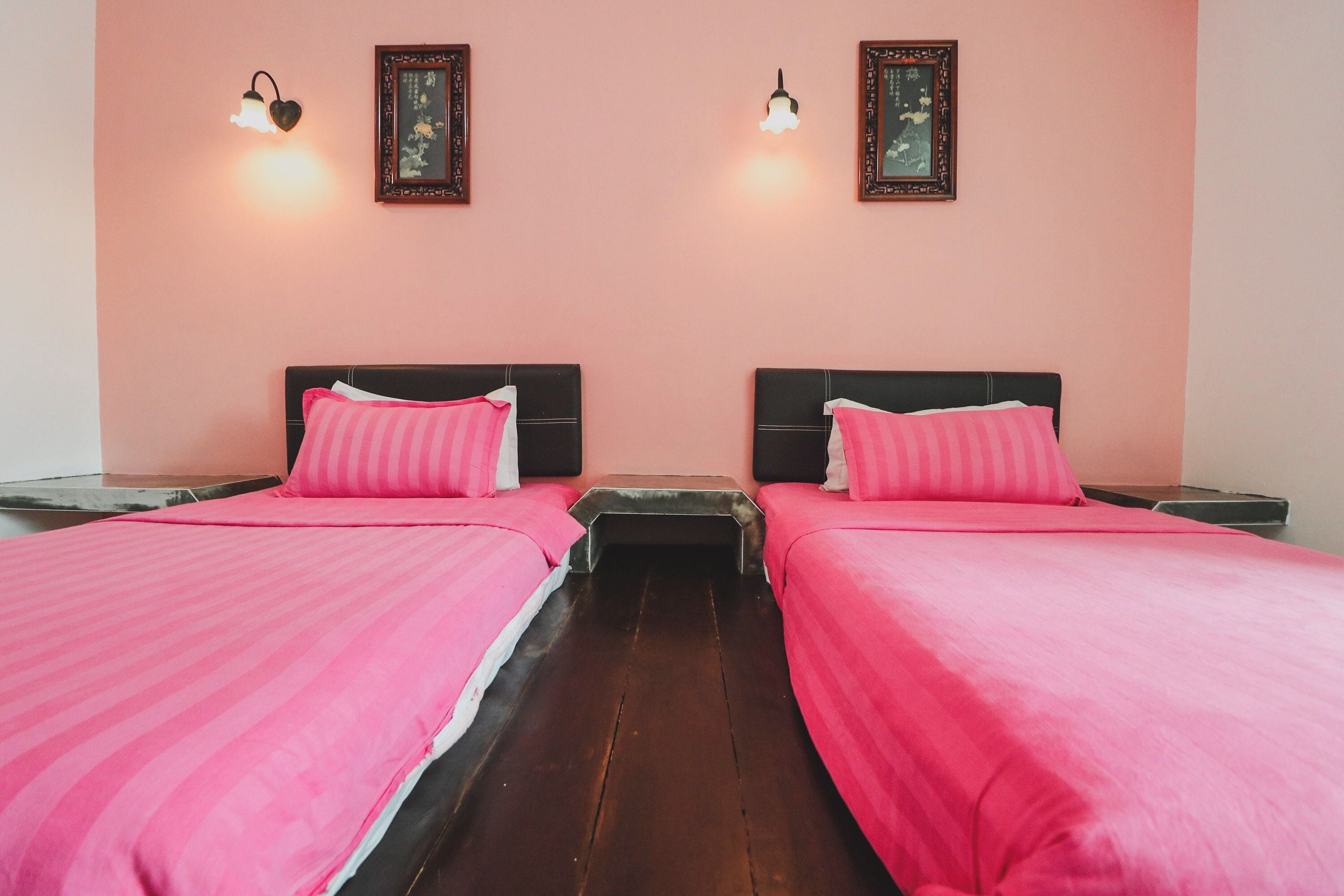 Una habitación con dos camas, cada una con una colcha rosa y almohadas a juego. Hay lámparas de pared y cuadros enmarcados sobre las camas.