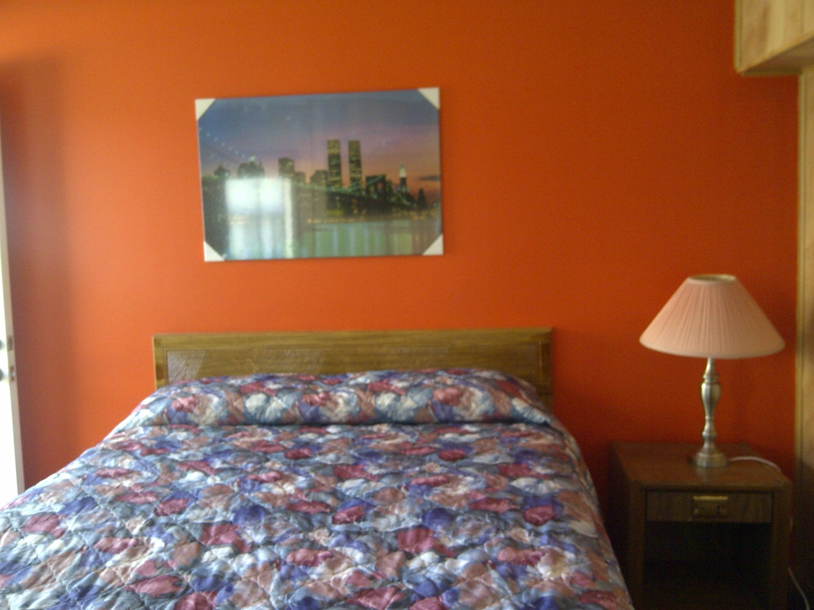 Una cama con colcha floral, una mesita de noche con lámpara y un cuadro de paisaje urbano en la pared.