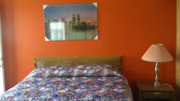 Una cama con colcha floral, una mesita de noche con lámpara y un cuadro de paisaje urbano en la pared.